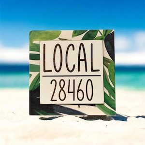 May include: A square wooden sign with the word "LOCAL" above the number "28460" in black lettering. The sign is set against a backdrop of green tropical leaves and a white background. The sign is on a sandy beach with a blue sky and ocean in the background.