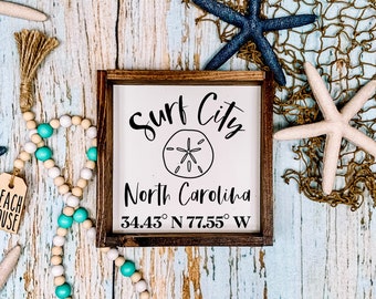 May include: A wooden sign with the text "Surf City North Carolina 34.43° N 77.55° W" and a sand dollar graphic. The sign is surrounded by beach decor, including a starfish, a string of beads, and a net.