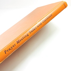 May include: An orange book titled "Prayer Meeting Talks and Outlines" with the word "BAKER" printed on the top edge. The book has a light peach-colored cover and is set against a white background.