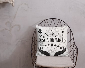 Könnte beinhalten: Ein weißes Kissen mit schwarz-weißen Illustrationen eines Nachtfalters, Mondphasen, eines Vogels und Händen, die eine schlafende Katze halten. Der Text "Just A Bit Witchy" ist in Schwarz auf dem Kissen gedruckt.