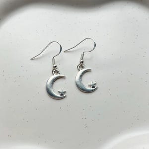 May include: A pair of silver-coloured earrings. Each earring features a crescent moon shape with a small star charm. The earrings have a simple hook design for pierced ears. The earrings are displayed on a white surface.