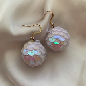 Puede incluir: Un par de pendientes colgantes blancos iridiscentes con ganchos dorados. Los pendientes están hechos de pequeñas lentejuelas redondas que se agrupan para formar una forma esférica.