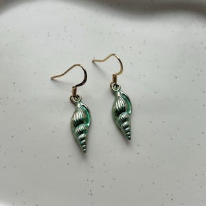 Puede incluir: Un par de pendientes de concha con ganchos dorados. Los pendientes son de color verde mar y tienen un diseño detallado y texturizado. Los pendientes están diseñados para colgar y son adecuados para el uso diario.