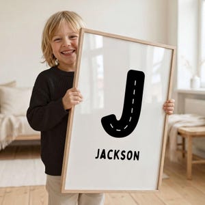 May include: A rectangular framed wall art print featuring a large black letter J designed to resemble a road with a white dashed centre line. Below the letter, the name JACKSON is printed in a bold, black, sans-serif font on a white background.
