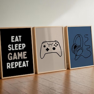 Peut inclure: Trois affiches encadrées. La première est noire avec le texte blanc EAT SLEEP GAME REPEAT. La deuxième est beige clair avec un dessin au trait noir d'une manette de jeu vidéo. La troisième est bleue avec un dessin au trait noir d'un casque filaire.