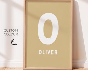 May include: A framed wall art print featuring a large white letter O centered on a solid tan background. Below the letter, the name OLIVER is printed in white capital letters. To the left of the frame, the text CUSTOM COLOUR is visible.