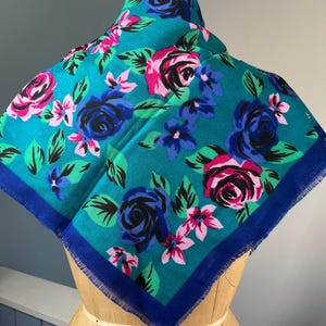 May include: A square scarf with a teal background and a floral pattern. The pattern features pink, blue, and black roses with green leaves. The scarf has a dark blue border and a fringed edge. The scarf is displayed on a mannequin.