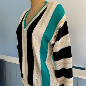 May include: A vintage V-neck sweater with a bold striped design. The sweater features alternating black, white, and teal stripes, embellished with shimmering sequins. The sleeves and neckline are trimmed in teal, creating a striking contrast.