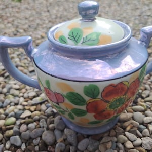 May include: A blue and white porcelain teapot with a floral design. The teapot has a lid with a knob on top and a handle on the side. The teapot is sitting on a bed of small stones.