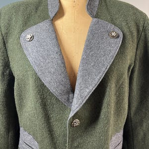 May include: A green wool jacket with gray accents. The jacket features a gray collar and lapels, with decorative silver buttons and leaf-shaped gray pocket details. The jacket is displayed on a mannequin.