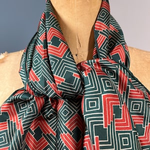 May include: A dark green scarf with a geometric pattern in red and white. The scarf is draped over a mannequin, showcasing the fabric's drape and design. The pattern consists of squares and lines.