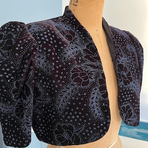 May include: A dark burgundy velvet bolero jacket with a floral pattern in light blue. The jacket features short, puffed sleeves and a high collar. The fabric has a soft sheen, and the jacket is displayed on a mannequin.