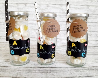 May include: Three glass bottles filled with colorful candies and topped with black and white striped straws. Each bottle has a brown paper tag with the text "Hurra! Endlich Schulkind" and a black chalkboard label with the letters "A, B, C" in yellow, blue, and pink.