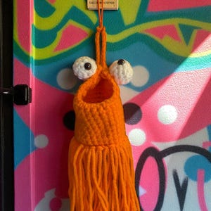 Könnte beinhalten: Ein handgefertigtes, orangefarbenes Häkelmonster mit weißen Wackelaugen und Fransen. Das Monster hängt an einem Holzanhänger mit dem Text "DALE & KPWNEY". Der Hintergrund ist ein farbenfrohes Graffiti-Design.