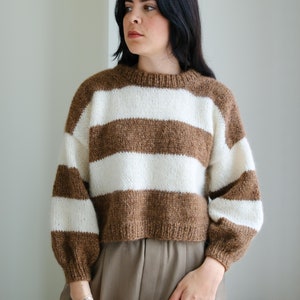 Può includere: Un maglione a righe marroni e bianche con un taglio corto e maniche lunghe. Il maglione è realizzato con un filato morbido e soffice.