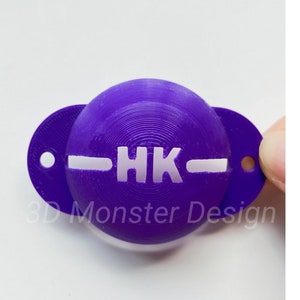 Può includere: Porta palline da golf in plastica viola con un logo "HK" bianco e una linea bianca al centro. Il porta palline è etichettato "3D Monster Design".