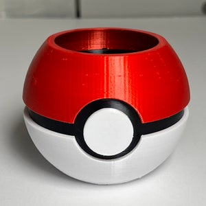 Può includere: Un vaso da fiori rosso e bianco a forma di Poké Ball con un bordo nero attorno al centro bianco. Il vaso è realizzato in plastica stampata in 3D.
