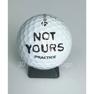 以下が含まれることがあります： 黒いスタンドの上に置かれた、黒い文字で「NOT YOURS PRACTICE」と書かれた白いゴルフボール