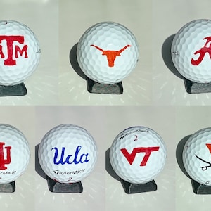 May include: Six white golf balls with different college logos painted on them. The logos are: Texas A&M, Texas Longhorns, Alabama, Indiana, UCLA, and Virginia Tech.