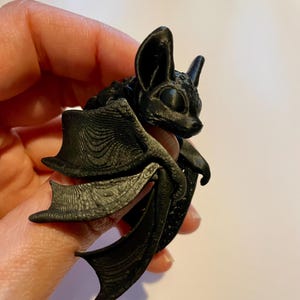 May include: A black 3D printed bat figurine with wings folded. The bat has large eyes and a detailed textured surface.