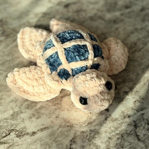 May include: A plush, handmade turtle toy. The turtle features a blue shell with a cream-colored grid pattern, cream-colored flippers, and a cream-colored head. The turtle has two black eyes.