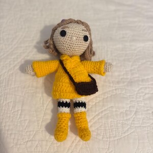 May include: A handmade crochet doll wearing a yellow dress, boots, and a brown purse. The doll has tan skin, brown hair, and black eyes. The doll's socks are white with black stripes. The doll is approximately 15 cm tall.