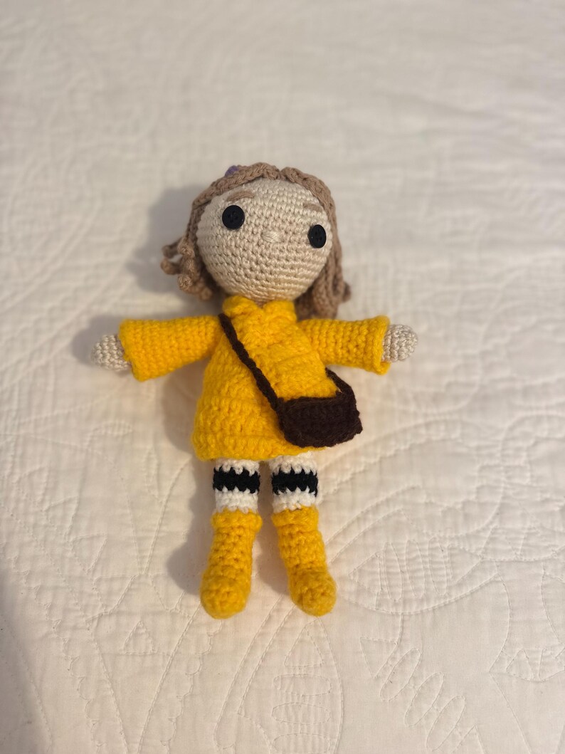 May include: A handmade crochet doll wearing a yellow dress, boots, and a brown purse. The doll has tan skin, brown hair, and black eyes. The doll's socks are white with black stripes. The doll is approximately 15 cm tall.