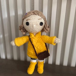 May include: A crocheted doll wearing a yellow coat, boots, and a brown satchel. The doll has light brown hair, black button eyes, and white arms and legs. A handcrafted, unique item.