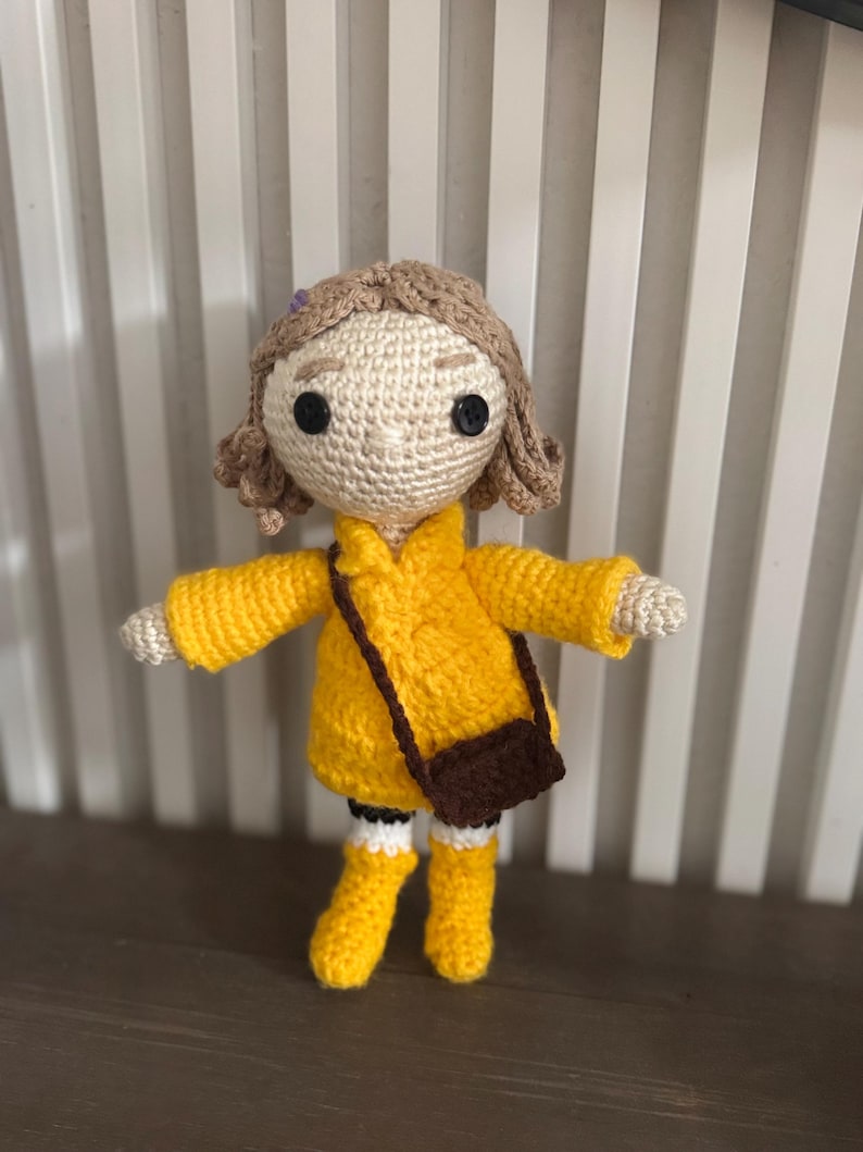 May include: A crocheted doll wearing a yellow coat, boots, and a brown satchel. The doll has light brown hair, black button eyes, and white arms and legs. A handcrafted, unique item.