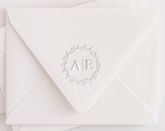 May include: Ivory-colored envelopes with a circular wreath design and the letters "AE" in the center. The envelopes are stacked, with one envelope in the foreground and others partially visible behind it. The design is simple and elegant.