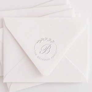 May include: Ivory-colored envelopes with a circular embossed design. The design includes a stylized letter "B" within a laurel wreath, with the words "THE BECKMAN FAMILY" encircling the design. The envelopes are stacked, with the top one in focus.