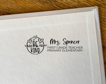 May include: A white paper with a black stamp that reads "BE KIND" inside a circle of hearts and swirls. Below the graphic, the text reads "Mrs. Spencer, First Grade Teacher, Primary Elementary."