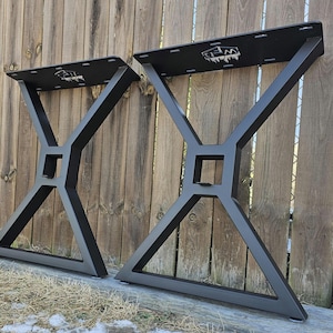 May include: Two black metal table legs with an X-shaped design. Each leg has a square center and a rectangular top with a logo. The legs are angled and appear sturdy, suitable for a modern table.