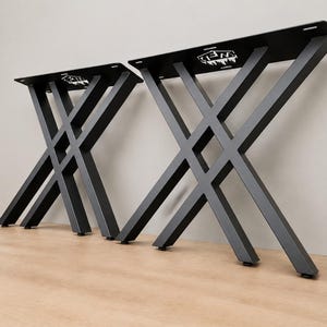 May include: Two black metal table legs featuring an X-shaped design with a flat rectangular mounting plate at the top. Each leg assembly includes a small cutout logo on the mounting plate. The legs have a matte finish and black floor protectors.