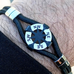 May include: A black leather bracelet with a compass charm featuring a black star and white compass points labeled NW, NE, SW, and SE.