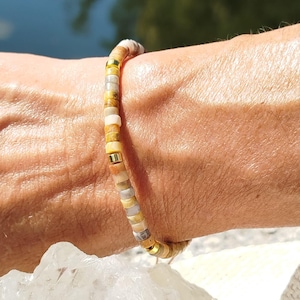 Op de afbeelding: Een kralenarmband met aardetinten, waaronder geel, bruin en crème. De armband is geregen op een dun koord en wordt om de pols gedragen. De kralen zijn klein en rond, wat een delicate en natuurlijke uitstraling creëert. Een sieraad.