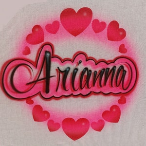 May include: A white t-shirt with the name "Ari'anna" written in black cursive letters surrounded by a circle of pink hearts.