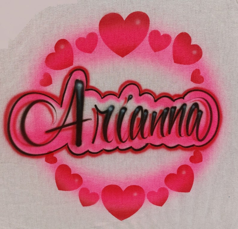 May include: A white t-shirt with the name "Ari'anna" written in black cursive letters surrounded by a circle of pink hearts.