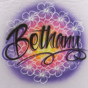 May include: A white t-shirt with a purple, pink, orange, and yellow gradient background. The background is decorated with a circle of white hibiscus flowers. The name "Bethany" is written in black cursive script over the flowers.