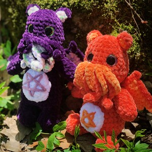 May include: Two crocheted plush toys, one purple with a pentagram and the other orange with a star. Both toys have large eyes and are made with a textured yarn.