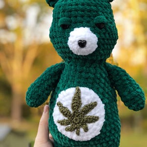 May include: A handmade, dark green plush bear with a white belly patch featuring a green cannabis leaf design. The bear has a white muzzle, black eyes, and a black heart-shaped nose. The bear is held against a blurred outdoor background.