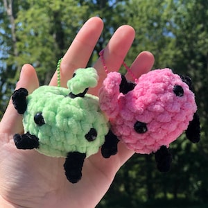 May include: Two crocheted plush toys, one green and one pink, with black eyes and legs. The toys are attached to keychains.