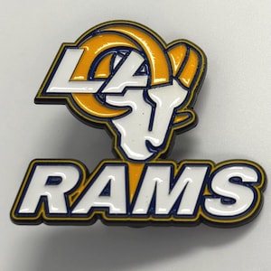 May include: A metal enamel pin featuring the Los Angeles Rams logo. The design includes a stylized white ram head with yellow horns, the letters LA in white, and the word RAMS in large white block letters, all outlined in dark blue and black.