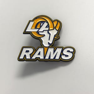 May include: A metal enamel pin featuring the Los Angeles Rams logo. The design includes a white ram head with yellow horns, the letters LA in white, and the word RAMS in white block letters, all outlined in dark blue against a white background.