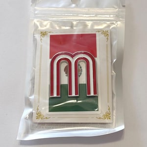 May include: A metal lapel pin shaped like the letter M with red and white stripes, mounted on a rectangular backing card featuring the green, white, and red colors of the Mexican flag with a central emblem, all enclosed in a clear plastic bag.