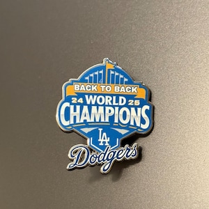 Może przedstawiać: Niebiesko-biała przypinka upamiętniająca zwycięstwa Los Angeles Dodgers w World Series. Przypinka zawiera napis "BACK TO BACK 24 WORLD 25 CHAMPIONS" oraz logo Dodgers.