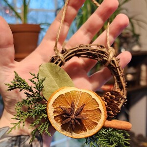 May include: A small, rustic wreath made of twigs and twine, decorated with dried orange slices, cinnamon sticks, star anise, pine cones, and evergreen sprigs.