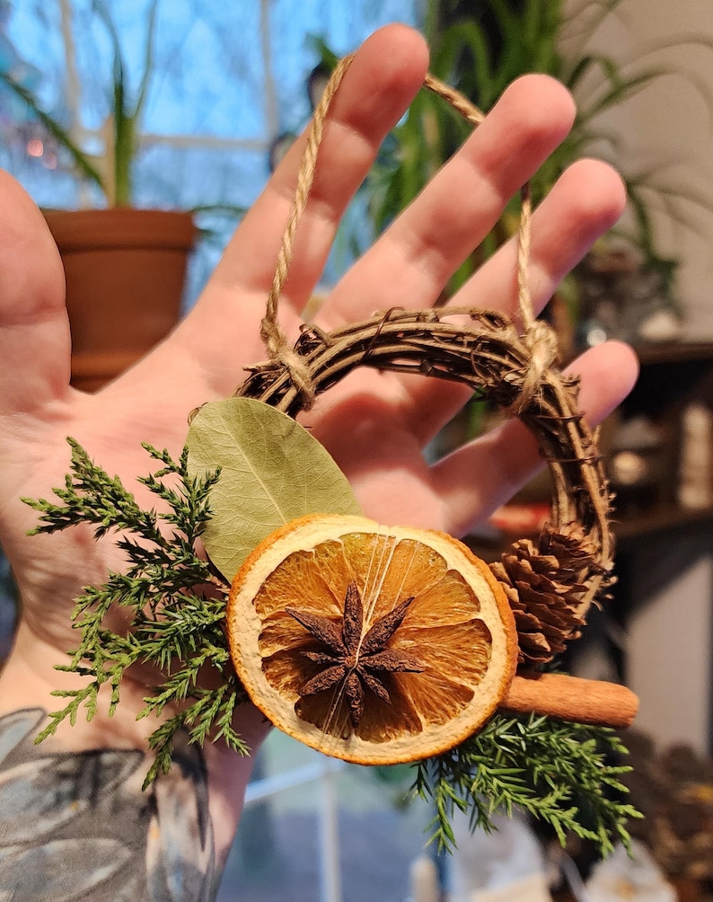 May include: A small, rustic wreath made of twigs and twine, decorated with dried orange slices, cinnamon sticks, star anise, pine cones, and evergreen sprigs.