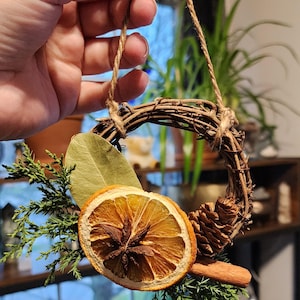 May include: A small, rustic wreath made of twigs, decorated with dried orange slices, cinnamon sticks, bay leaves, pine cones, and evergreen sprigs. The wreath is hung with twine.