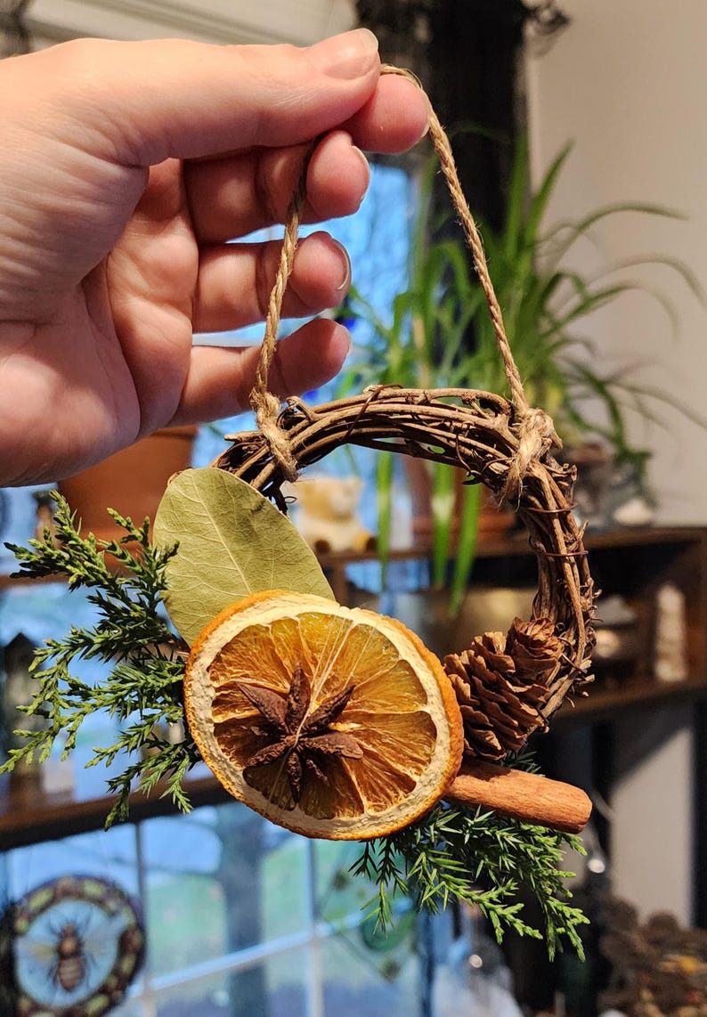May include: A small, rustic wreath made of twigs, decorated with dried orange slices, cinnamon sticks, bay leaves, pine cones, and evergreen sprigs. The wreath is hung with twine.